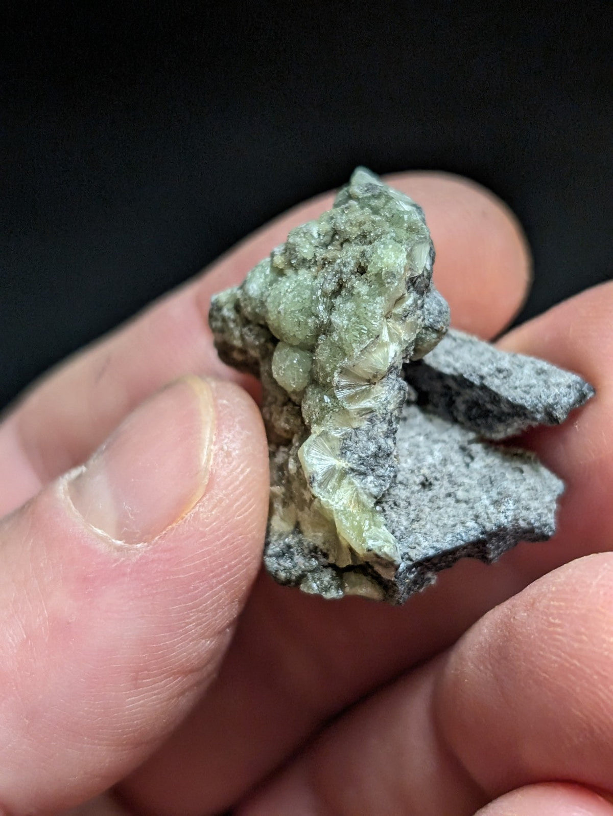 A hand holds a rough, irregularly shaped mineral specimen featuring translucent green crystals embedded in a dark gray matrix.