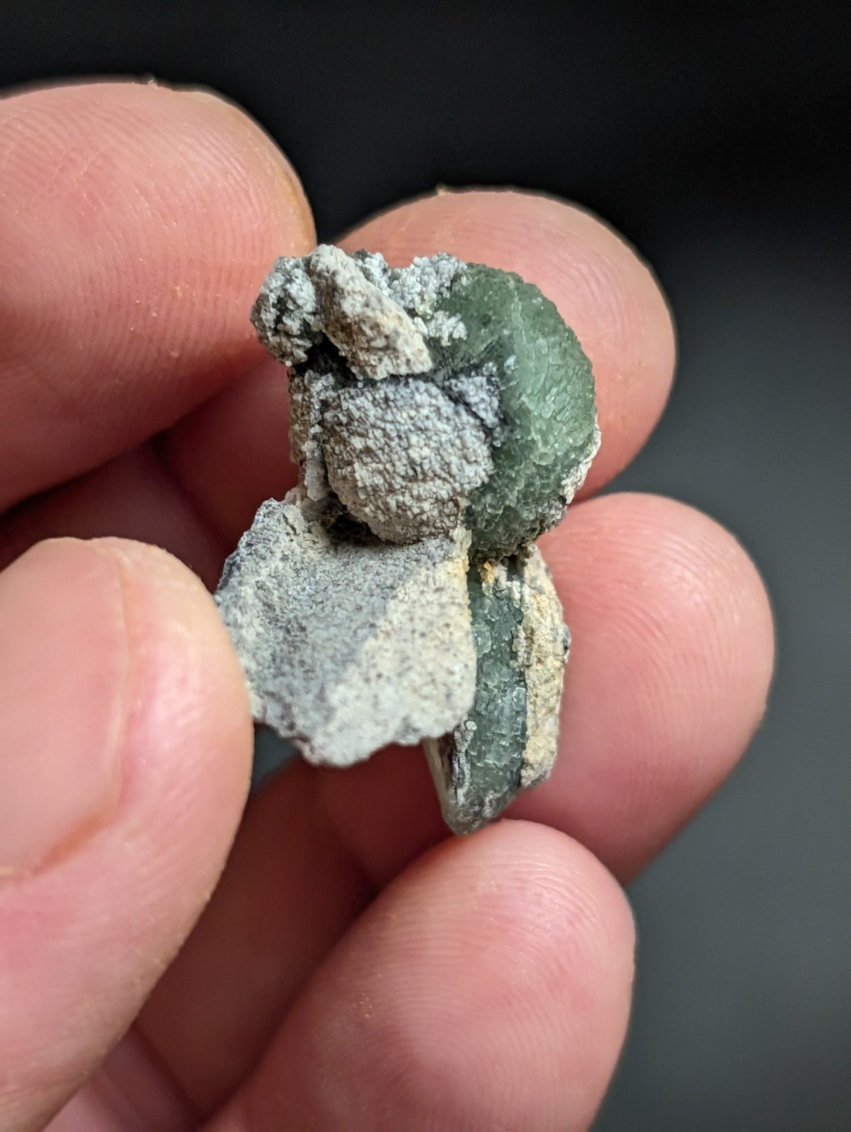 A green, rough-textured mineral specimen with grayish-white crusty patches and a jagged, irregular shape.