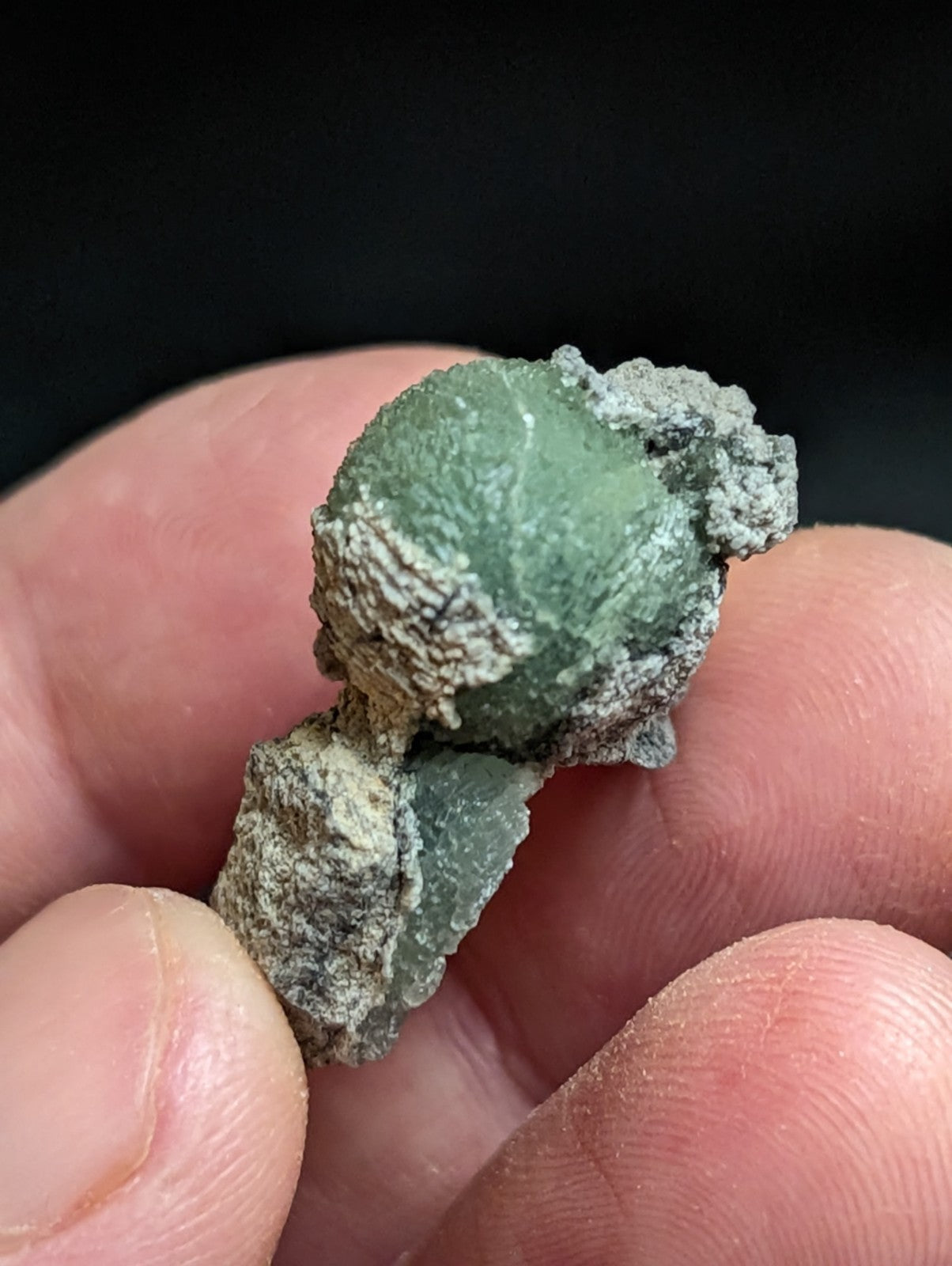 A rough, greenish mineral specimen with a textured surface and grayish crusty patches.