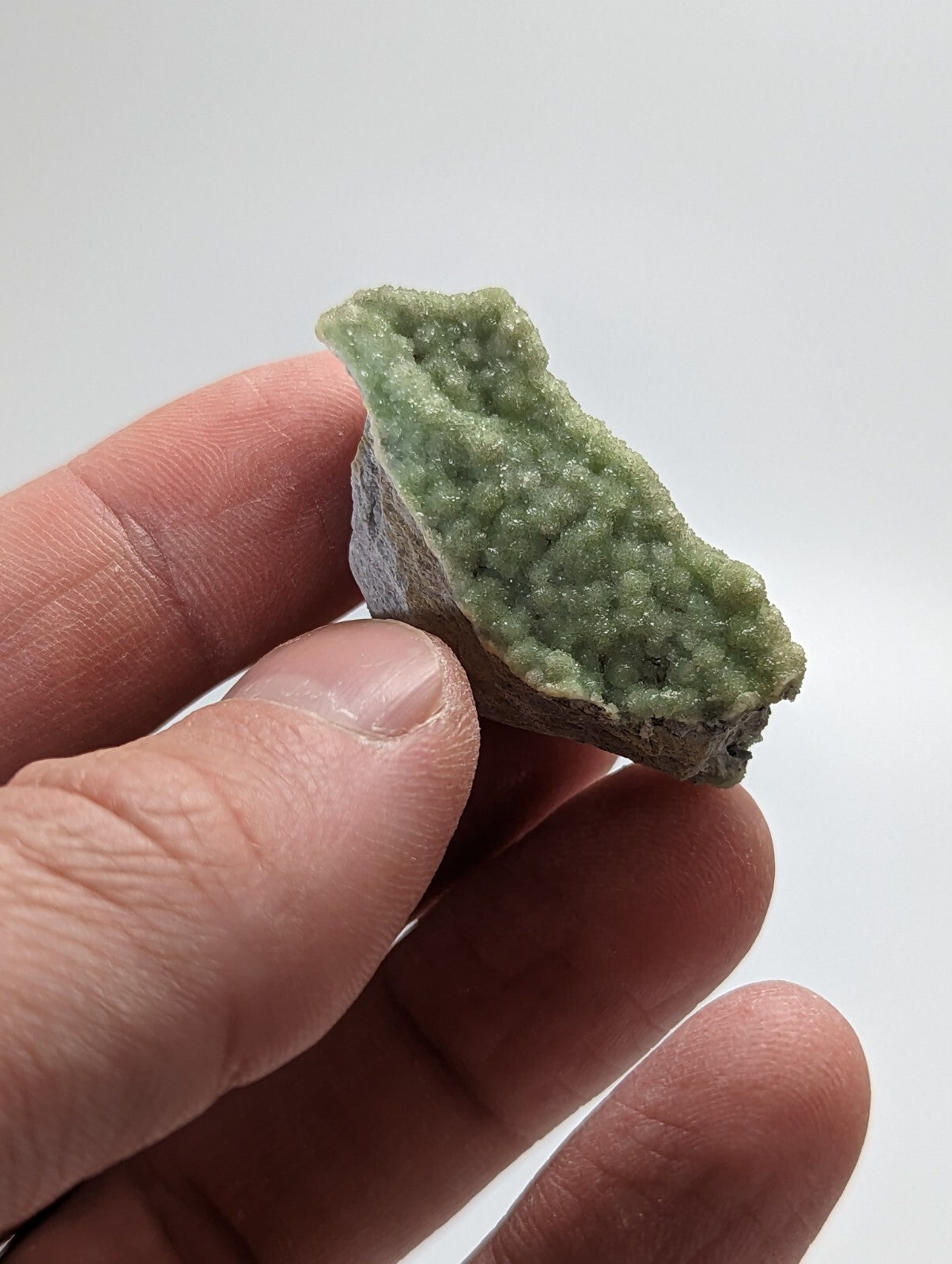A hand holds a small, irregularly shaped green mineral specimen with a textured surface covered in tiny crystalline formations.