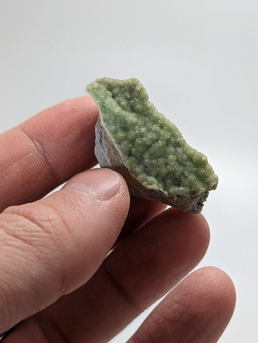 A hand holds a small, irregularly shaped green mineral specimen with a textured surface covered in tiny crystalline formations.