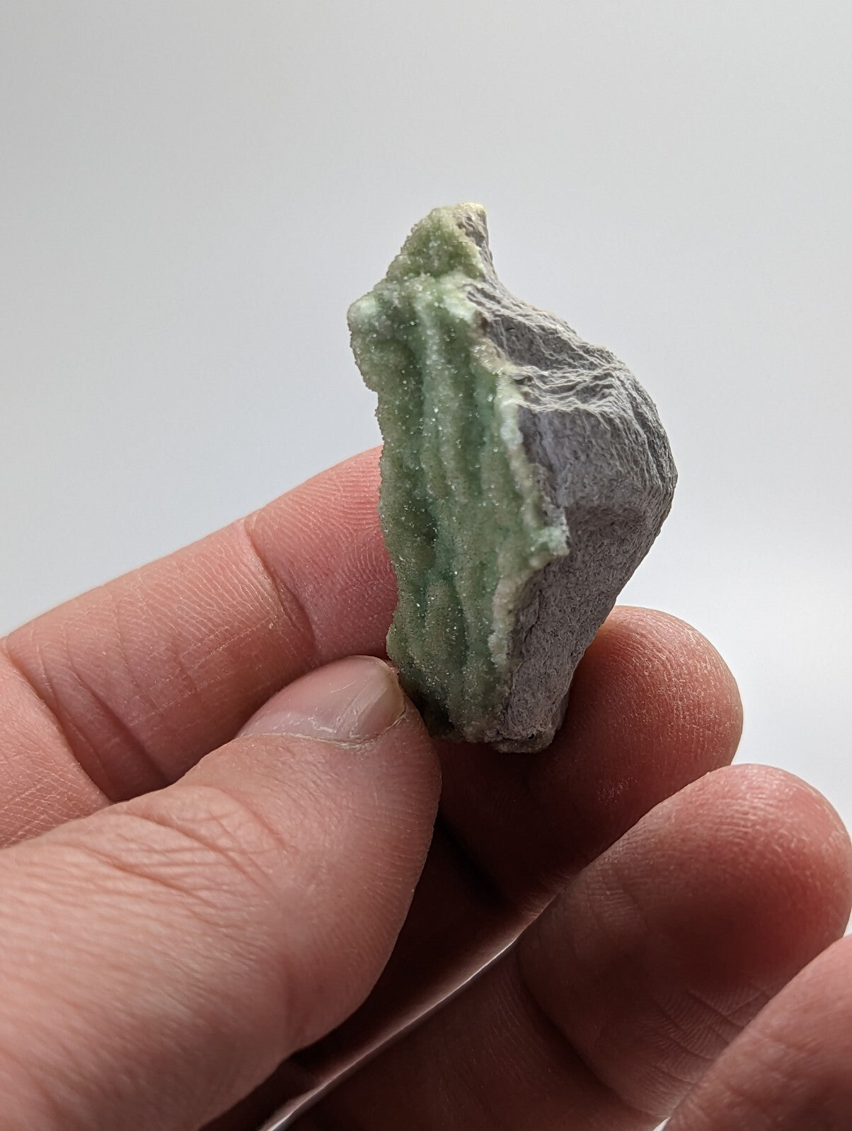 A hand holds a rough, irregularly shaped mineral specimen with a pale green crystalline growth emerging from a darker gray rock matrix.