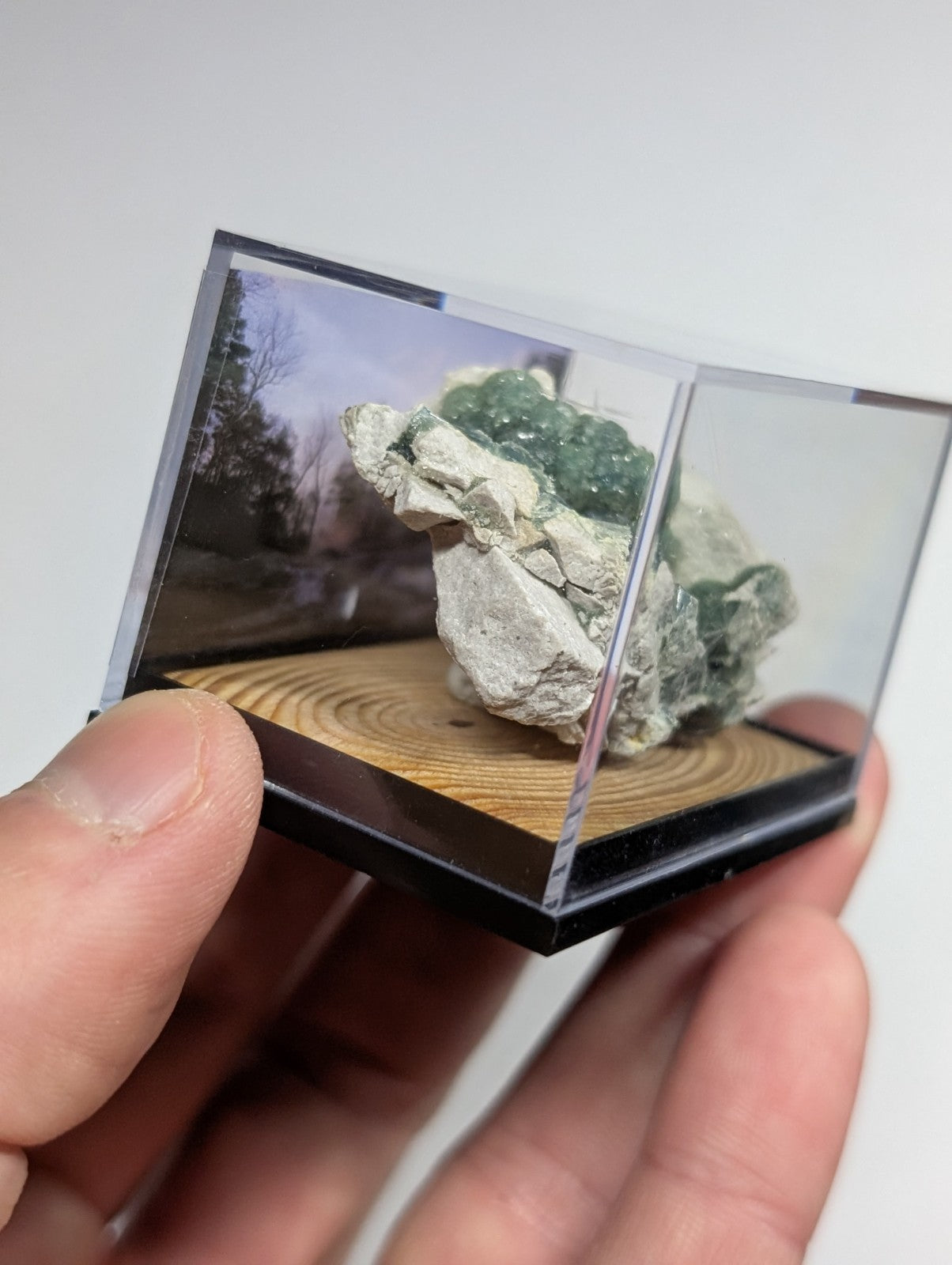 A miniature clear acrylic display case holding a green and white mineral specimen on a wooden base.