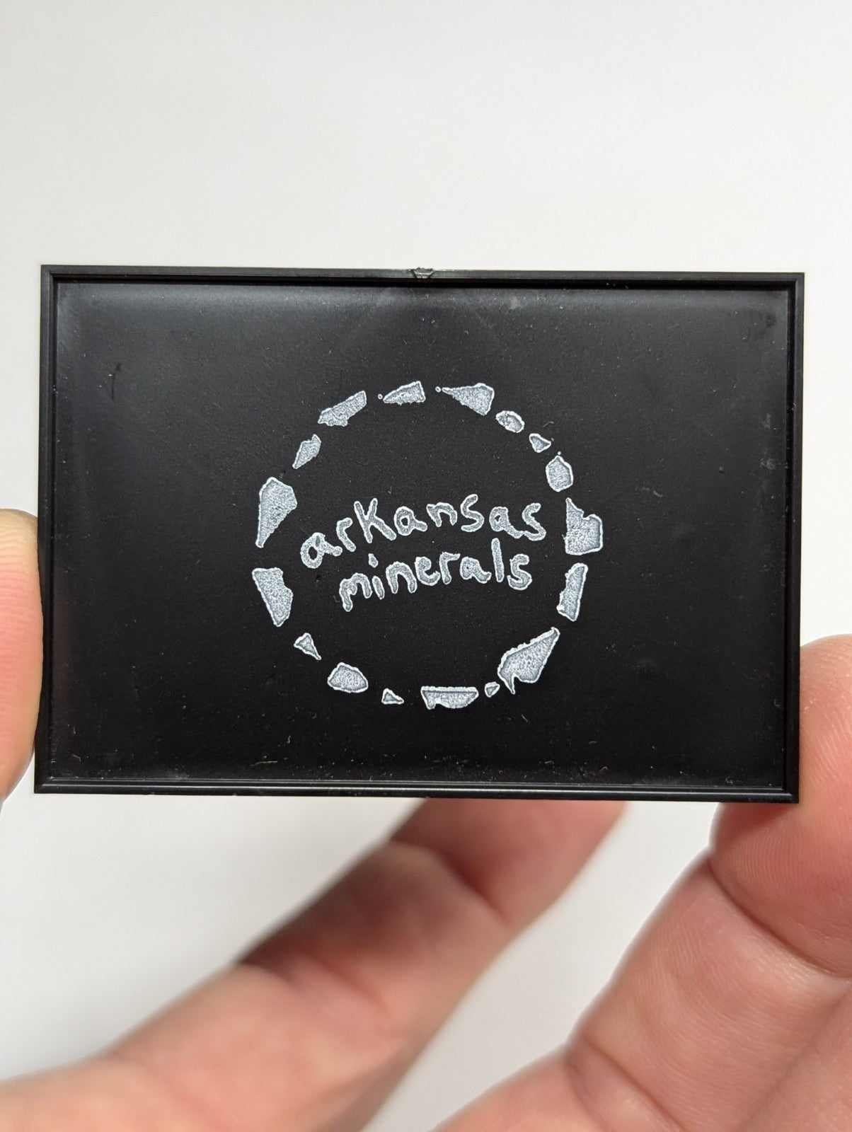 Black rectangular mineral sample holder with silver engraved ’arkansas minerals’ logo surrounded by a circle of stylized rock fragments.