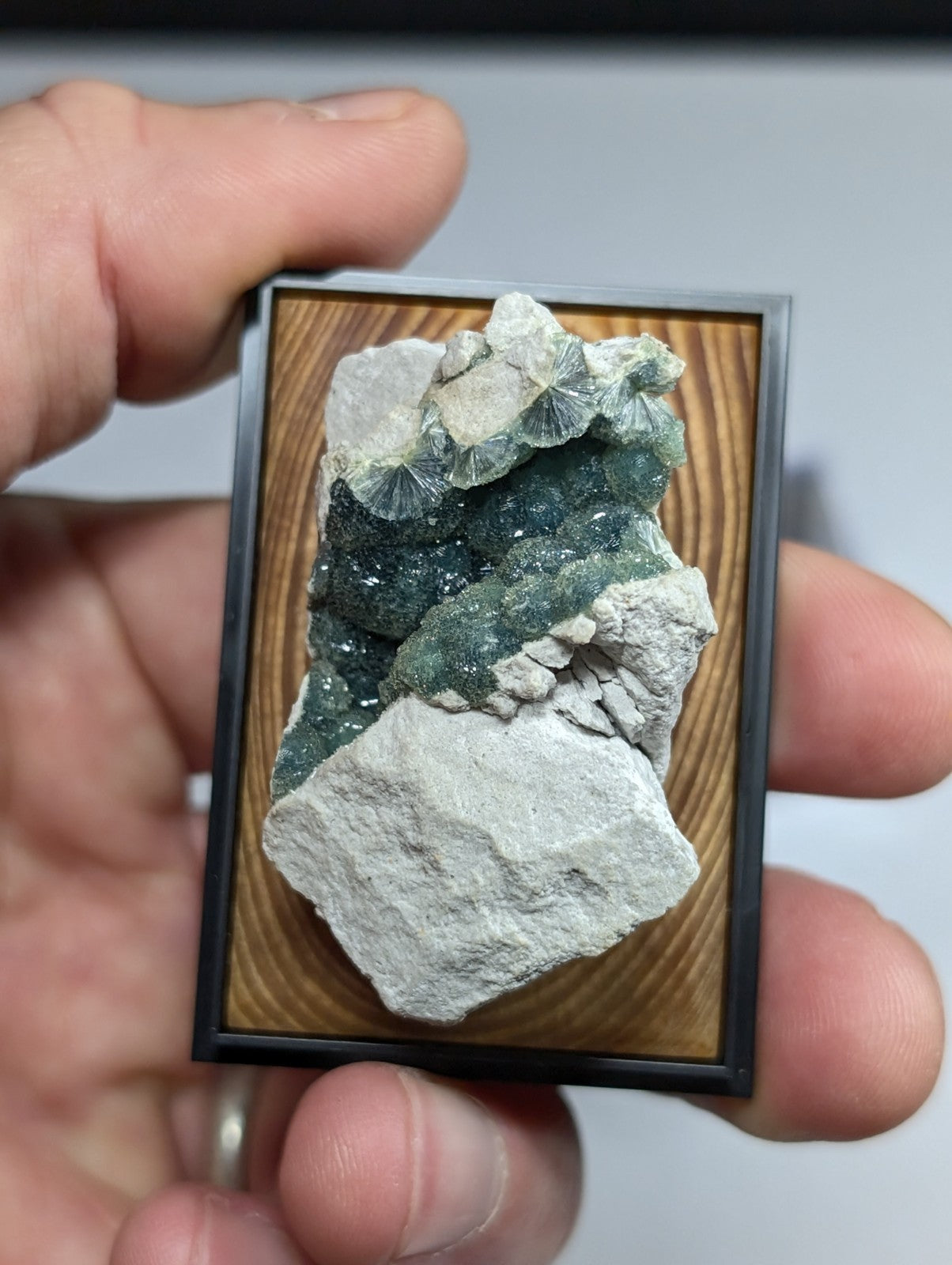 A framed mineral specimen featuring a cluster of green and white crystals with fan-shaped formations set against a light wood-grain background.