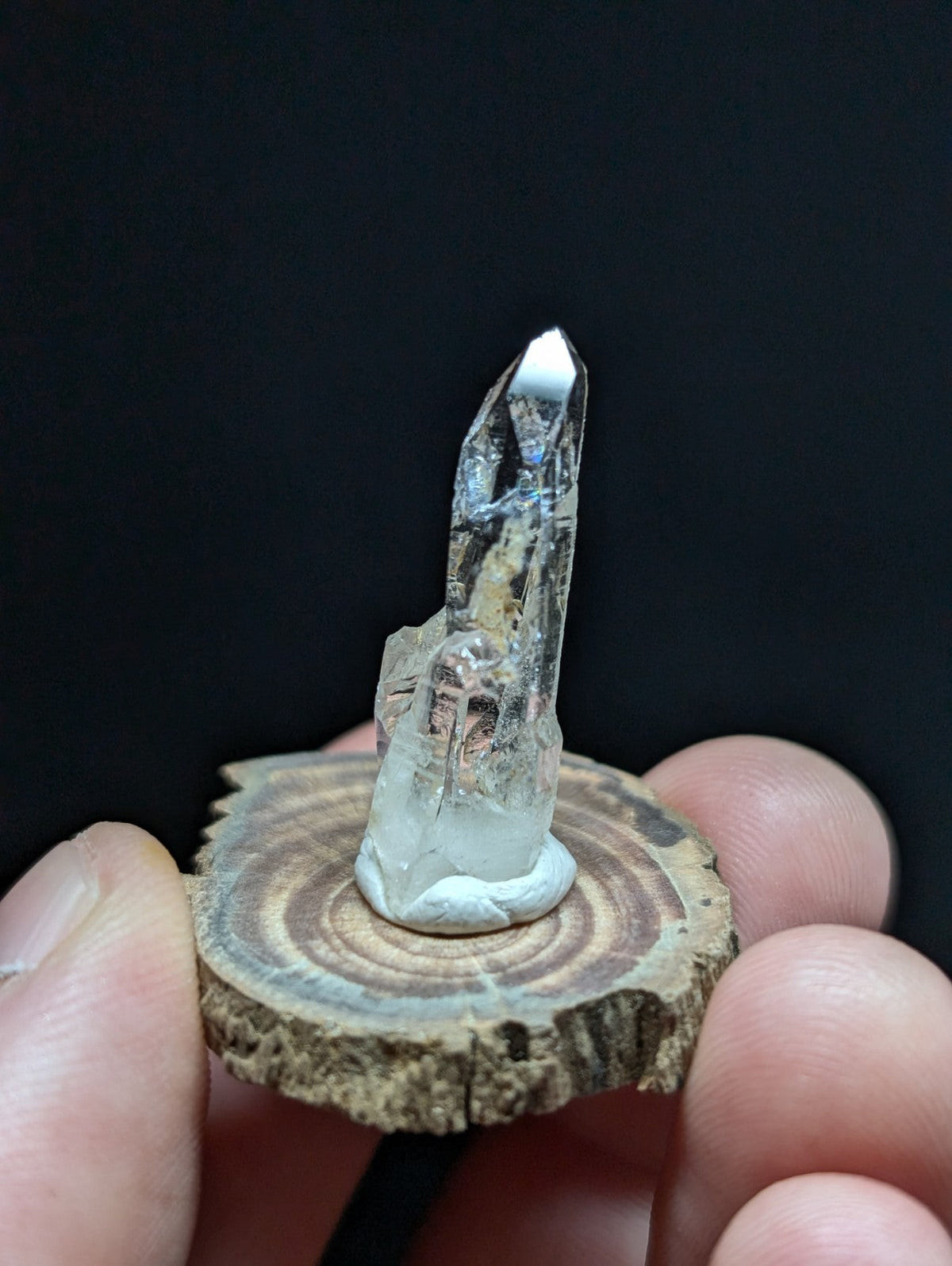 A clear quartz crystal with a pointed tip and natural inclusions, mounted on a rustic wooden slice.