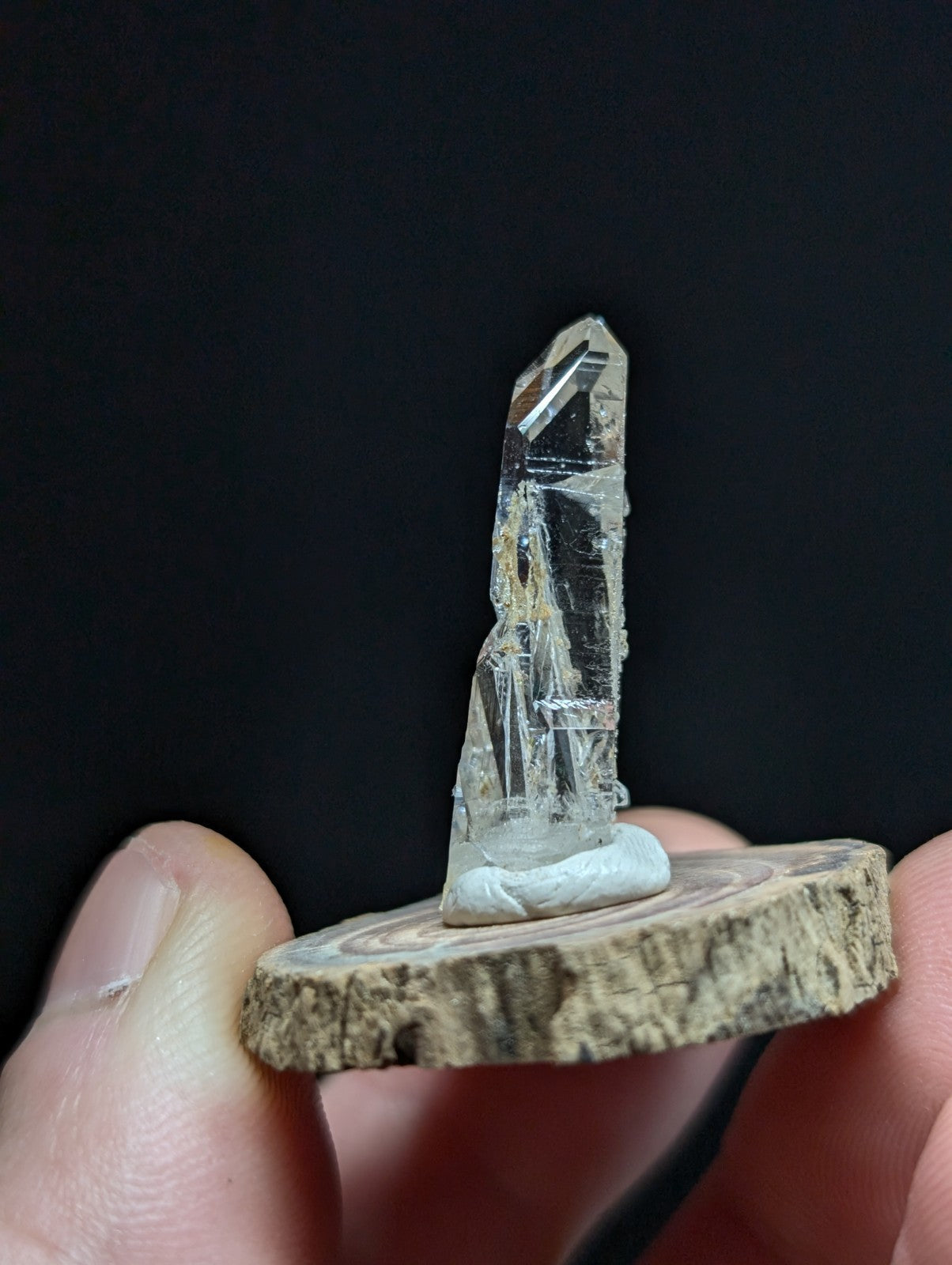 A clear quartz crystal with a pointed tip and natural facets, mounted on a rustic wooden slice.