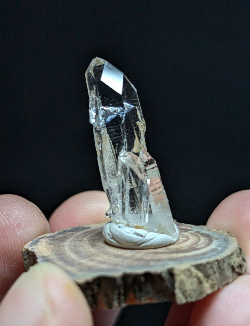 A clear, faceted quartz crystal with a pointed tip and natural imperfections sits atop a rustic wooden slice.
